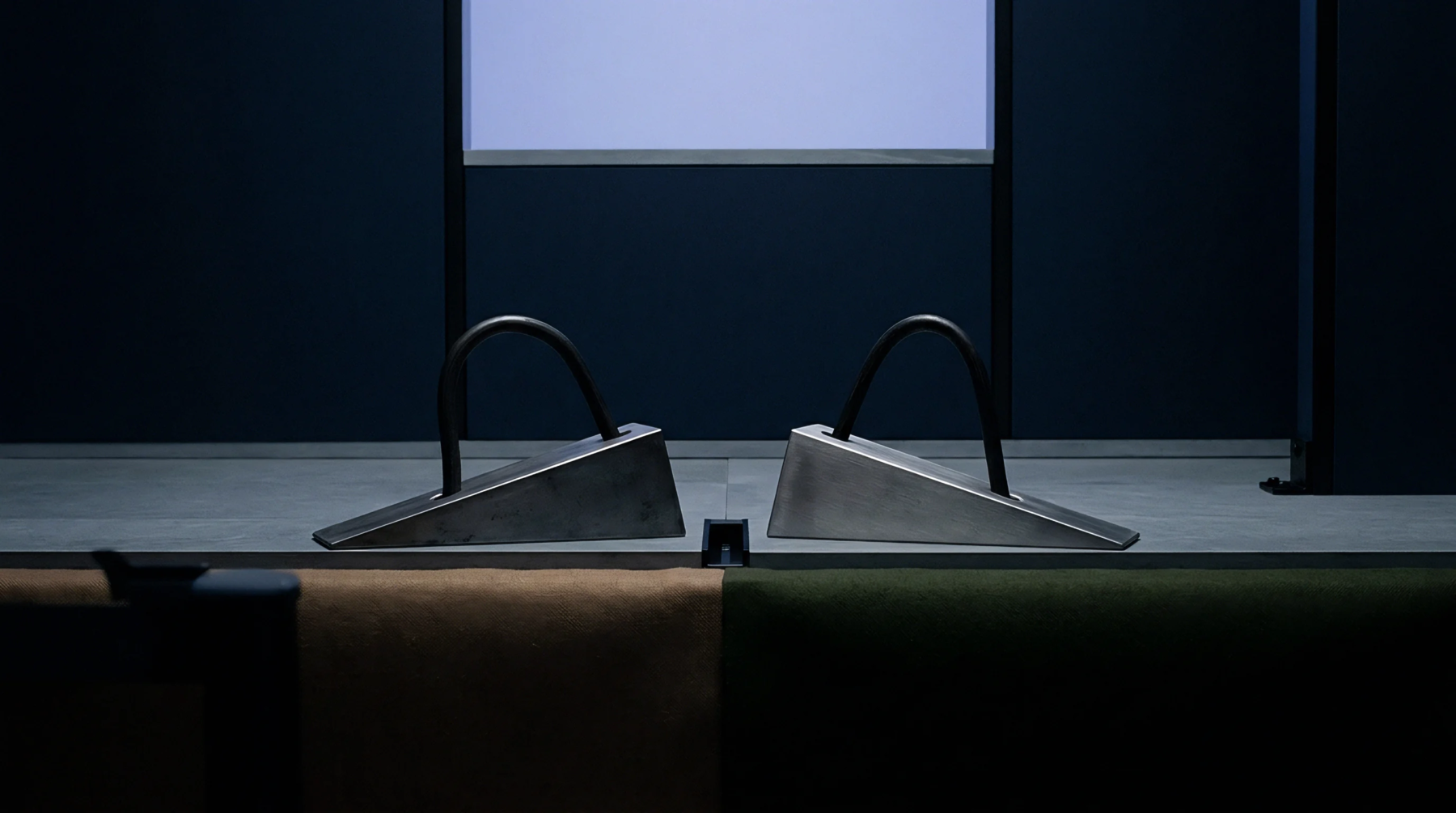Two brushed steel wedge fixtures featuring black tubular handles sit symmetrically on a grey workshop platform before a dark partition.